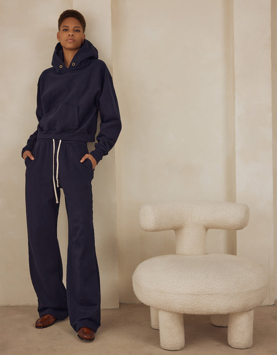 Text on image says "Shop Hoodies Made For Her" - In image of a woman standing in a navy cropped hoodie with gold eyelets and navy puddle pants with drawstrings, standing next to a fuzzy ivory modern seat.