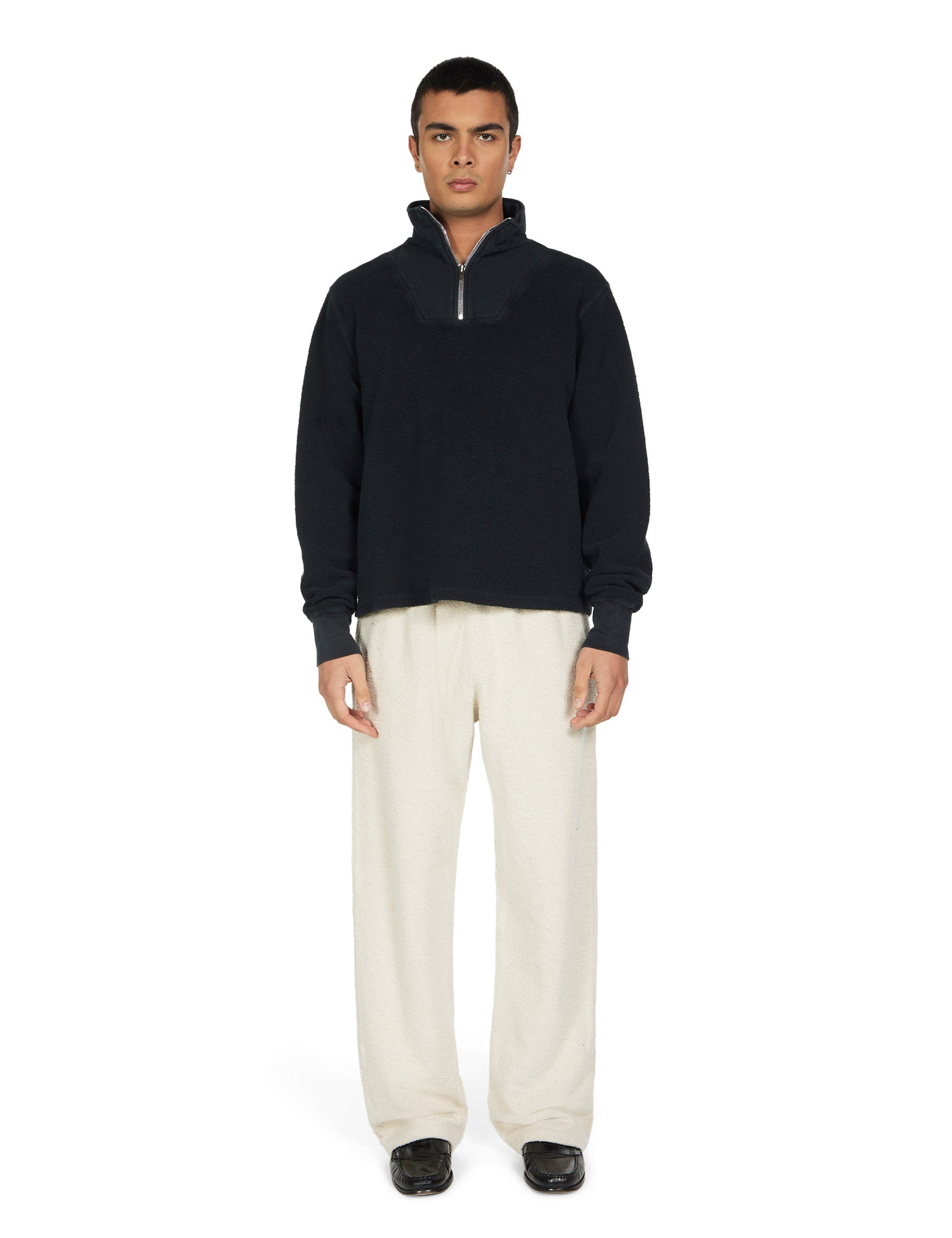 Man wearing a black half-zip sweatshirt and ivory pants on a white background