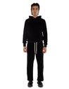 A man wearing a black velour hoodie with gold eyelets, black pants with white drawstrings, and black boots on a white background