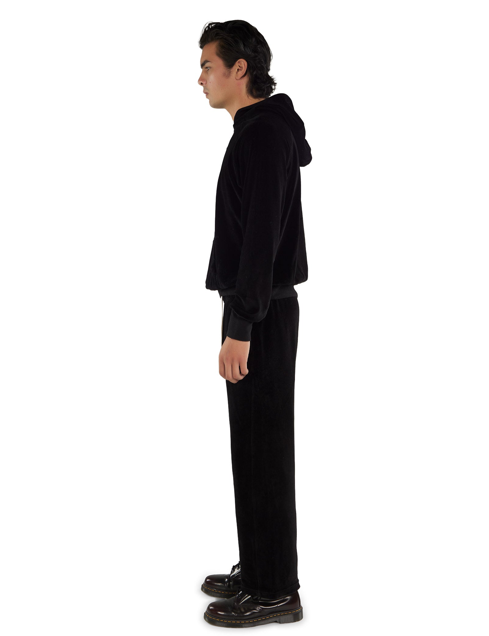 A side view of a man wearing a black velour hoodie with gold eyelets, black pants with white drawstrings, and black boots on a white background