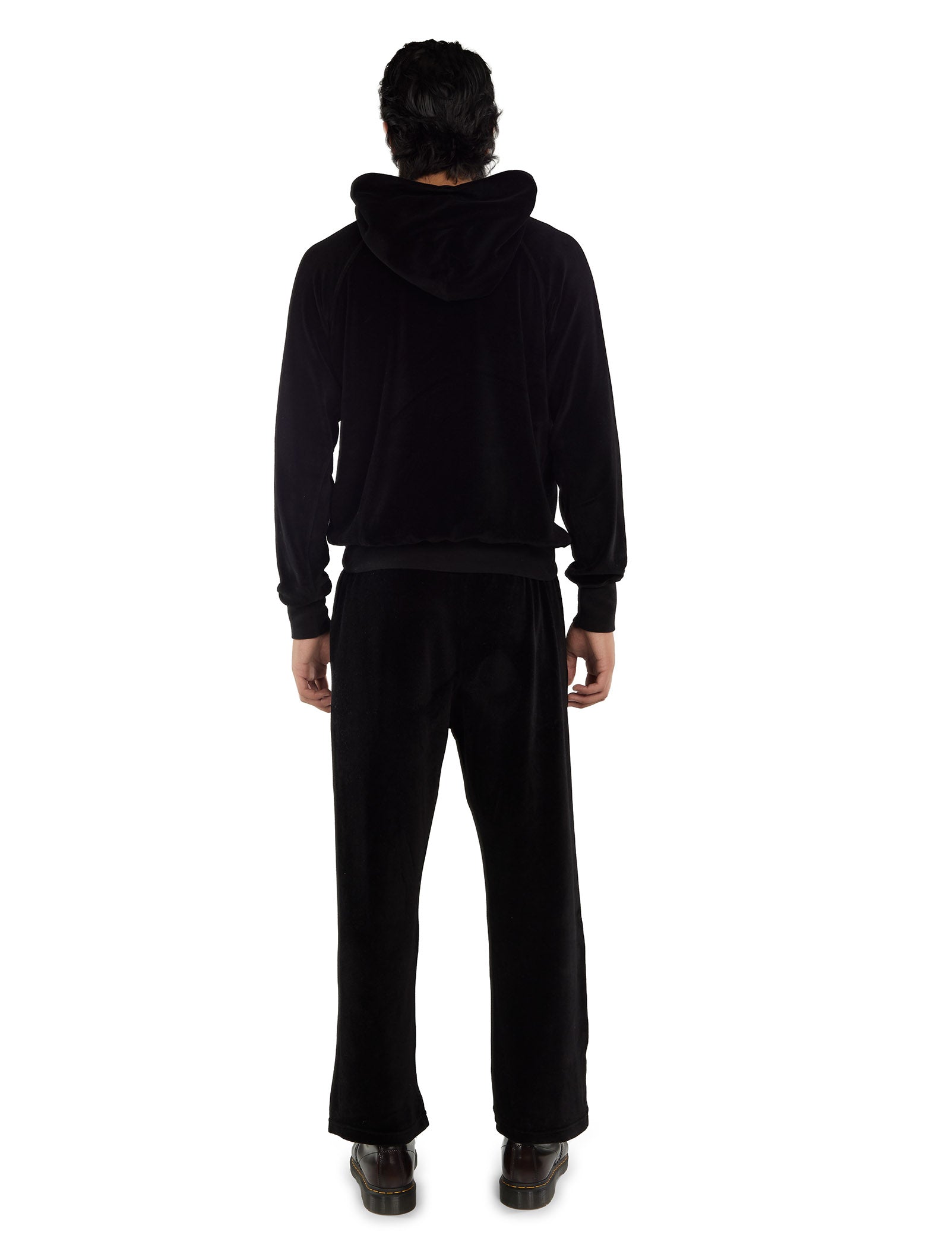 A back view of a man wearing a black velour hoodie with gold eyelets, black pants with white drawstrings, and black boots on a white background