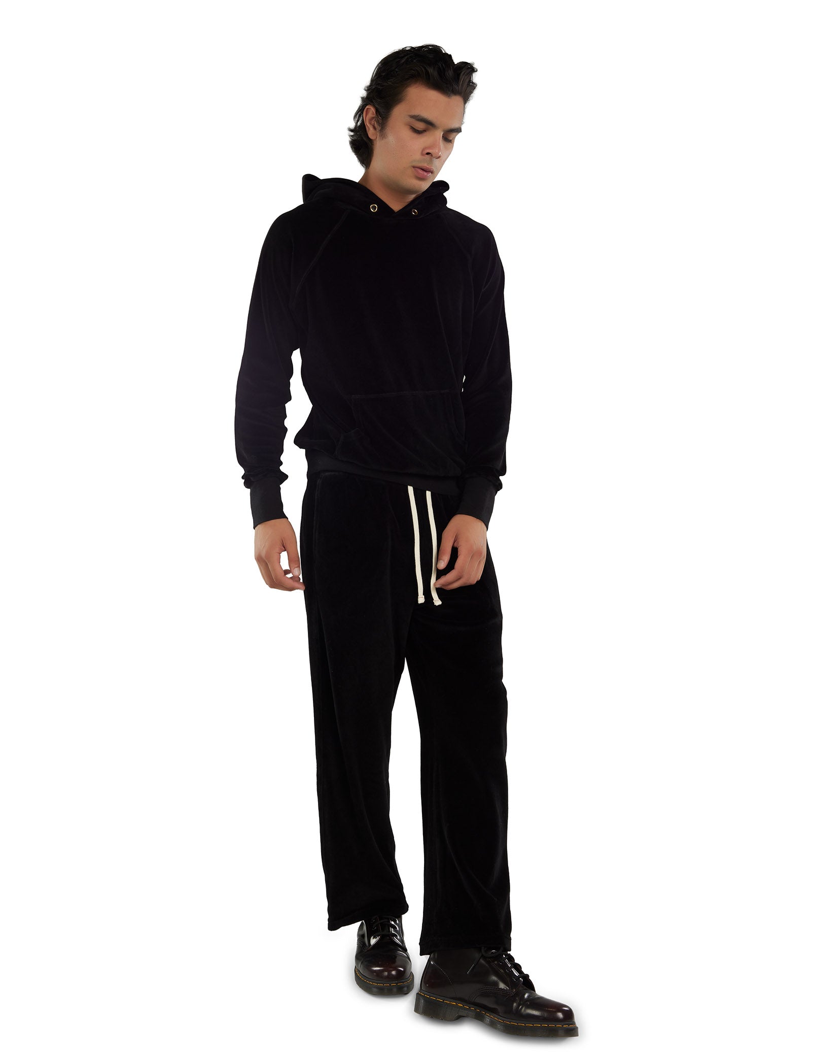 A man wearing a black velour hoodie with gold eyelets, black pants with white drawstrings, and black boots on a white background